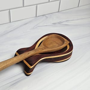 May include: A wooden spoon resting on a contoured spoon rest. The spoon rest features alternating layers of dark brown and light tan wood, creating a striped pattern. The spoon is crafted from a single piece of light brown wood with a visible grain.