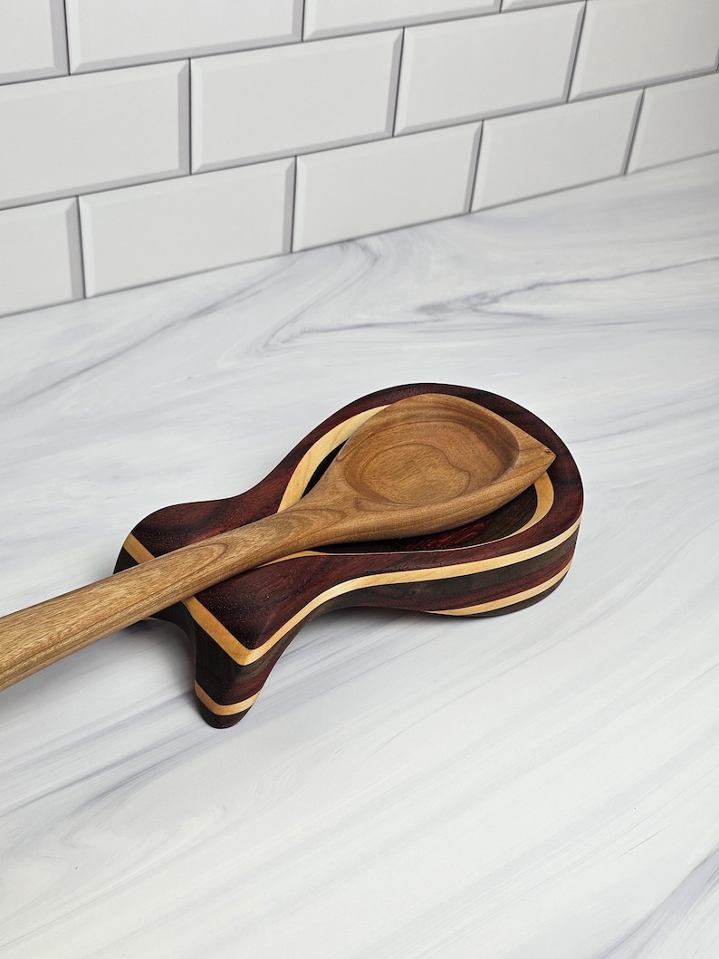 May include: A wooden spoon resting on a contoured spoon rest. The spoon rest features alternating layers of dark brown and light tan wood, creating a striped pattern. The spoon is crafted from a single piece of light brown wood with a visible grain.