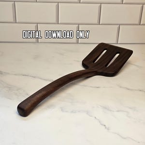 May include: A dark brown wooden spatula with a curved handle and two vertical slots in the head. The text DIGITAL DOWNLOAD ONLY is visible in the background above the spatula, which rests on a white surface with grey veining.