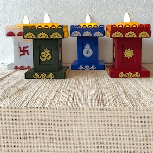 以下が含まれることがあります： LEDの炎のライトが付いた、カラフルな装飾用キャンドルホルダー4つ。キャンドルホルダーは白、緑、青、赤で、それぞれユニークなシンボルが付いています。ベージュの織り込まれた表面に置かれています。