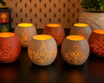 May include: A collection of seven spherical terracotta candle holders in shades of red and beige. Each holder features intricate cut-out designs including peacocks, floral motifs, and geometric patterns, illuminated from within by warm light.