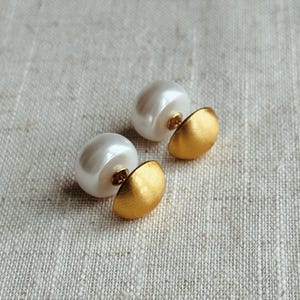 Könnte beinhalten: Ein Paar elegante Ohrringe mit großen, glänzenden weißen Perlen, kombiniert mit goldfarbenen, halbmondförmigen Akzenten. Die Ohrringe sind vor einem neutralen, strukturierten Hintergrund platziert, was ihr schlichtes, aber raffiniertes Design hervorhebt.