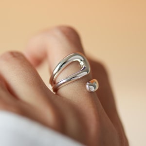 May include: A polished silver ring featuring an open, fluid, and organic shape. The band consists of a smooth, continuous metal loop that curves into a teardrop-like opening, ending in a small, rounded, and reflective silver bead detail.
