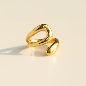 Puede incluir: Un anillo de tono dorado con un diseño abierto, fluido y asimétrico de superficies lisas y pulidas. La banda se curva en una forma continua y en bucle que crea un centro hueco, mostrando un acabado metálico reflectante en todo el objeto.