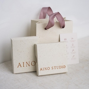 Könnte beinhalten: Eine cremefarbene Geschenktüte mit mauvefarbenen Bandgriffen, zwei passende cremefarbene Schachteln mit gesprenkelter Textur und eine kleine Informationskarte. Die Schachteln tragen den Text AINO und AINO STUDIO in kupferfarbener Schrift.