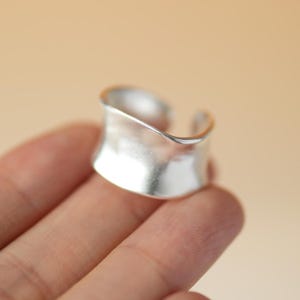 Könnte beinhalten: Ein breiter, offener Silberring mit einer glatten, matten Oberfläche. Das Band weist ein geschwungenes, konkaves Design auf, das sich zur Mitte hin leicht verjüngt und eine fließende, durchgehende Form bildet, die den Finger mit einer Öffnung umschließt.