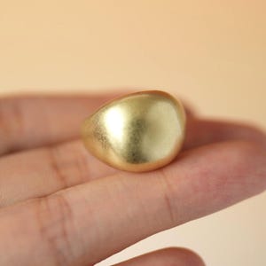 May include: A gold-toned ring featuring a smooth, rounded, and domed surface with a matte finish. The ring has a thick, solid band design that curves gently, reflecting light across its polished, convex top surface in a simple, circular form.