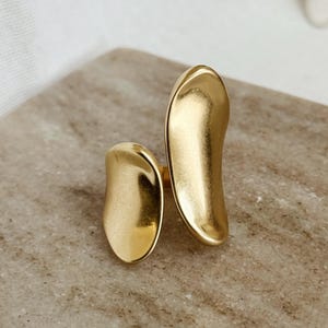 May include: A gold ring with an abstract design. The ring features two asymmetrical, elongated shapes that curve upwards. The ring is set against a neutral, textured background.