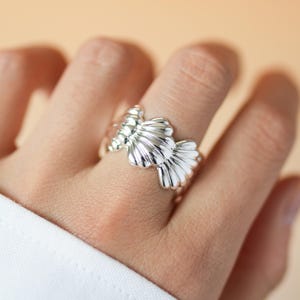 Puede incluir: Un anillo de plata pulida que presenta un diseño repetitivo de conchas marinas tridimensionales en forma de abanico. La banda metálica está elaborada con un acabado liso y reflectante, mostrando las crestas y bordes curvos definidos.