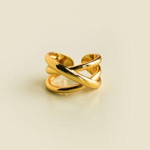 Pode incluir: Um anel dourado polido com um design cruzado aberto, apresentando duas bandas lisas e arredondadas que se cruzam no centro. O anel tem um acabamento metálico reflexivo e uma abertura larga de estilo ajustável na parte de trás.