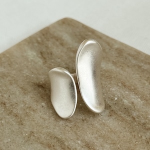 May include: A silver ring featuring two abstract, elongated forms. The ring has a matte finish and is set against a neutral backdrop. The design is modern and minimalist, suitable for everyday wear.