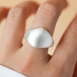 May include: A silver-toned ring featuring a smooth, rounded, and slightly asymmetrical top surface with a brushed matte finish. The band is solid and wraps around the finger, showcasing a clean, polished look with no additional engravings or stones.