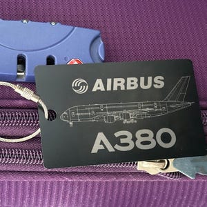 以下が含まれることがあります： エアバスのロゴとA380航空機のイラストが描かれた黒いラゲッジタグ。紫色のスーツケースに、青いダイヤルロックと一緒に取り付けられています。