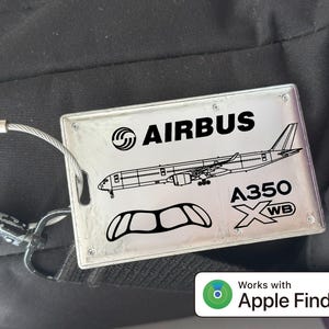 May include: Silver rectangular luggage tag with the Airbus logo, an airplane illustration, and the text "A350 XWB." The tag is attached to a bag with a metal wire loop. The tag also has the text "Works with Apple Find My."