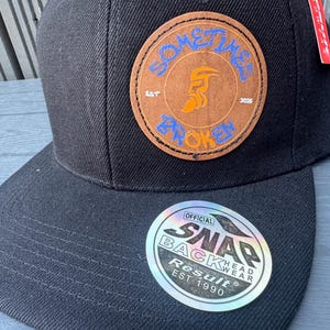 May include: Black snapback cap with a circular patch displaying the words "SOMETIMES BROKEN" in blue and an orange figure. The cap also has a "SNAP BACK" sticker.