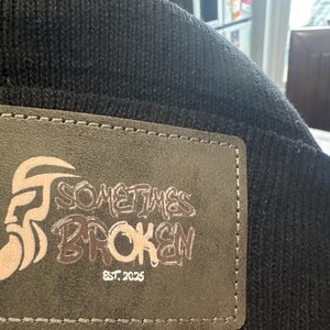 Op de afbeelding: Zwarte gebreide beanie met een rechthoekige leren patch. De patch toont een gestileerde afbeelding van een gezicht en de woorden "SOMETIMES BROKEN" en "EST. 2025" in een verweerde lettertype.
