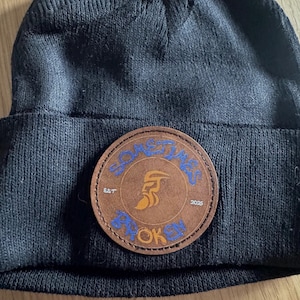Op de afbeelding: Zwarte beanie met een bruine ronde patch. De patch heeft de tekst "SOMETIMES BROKEN OK" in blauwe letters, met een oranje vlamontwerp. De beanie is gemaakt van zacht, gebreid materiaal. De patch heeft ook de tekst "EST 2025".