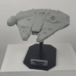 Può includere: Un modello grigio del Millennium Falcon, un'astronave immaginaria del franchise di Star Wars, è esposto su un supporto nero. Il modello è dettagliato con vari pannelli e caratteristiche, e il supporto ha un'etichetta che recita "Millennium Falcon."