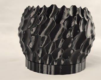Puede incluir: Una maceta negra texturizada con un diseño orgánico único. La superficie tiene un patrón de formas irregulares en relieve, lo que le da una apariencia rugosa y táctil. La maceta tiene una base ancha y un borde festoneado, adecuada para uso interior o exterior.