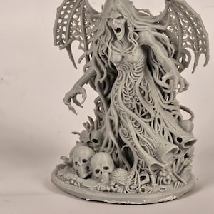 May include: A gray figurine of a winged creature with long hair and an open mouth, standing amidst skulls and organic shapes. The figure has detailed wings and a flowing dress-like form, with a menacing expression.