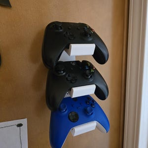 May include: A wall-mounted game controller holder with three controllers. Two black controllers are stacked above a blue controller. The controllers are held in place by white brackets, creating a space-saving storage solution for gaming accessories.