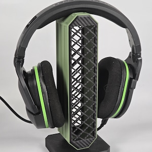 May include: Black and green gaming headphones on a green and black headphone stand. The stand features a diamond pattern and a black base. The headphones have green detailing.