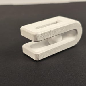 Könnte beinhalten: Ein weißes, 3D-gedrucktes Objekt mit U-förmigem Design. Das Objekt hat eine rechteckige Oberseite mit einem Schlitz und eine abgerundete Unterseite. Die Textur erscheint rau. Das Objekt ist wahrscheinlich ein Clip oder Halter.
