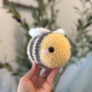 May include: A plush bee toy with a yellow and black striped body, white wings, and a small pink blush on its cheeks. The bee has a fuzzy texture and a black bead for an eye.