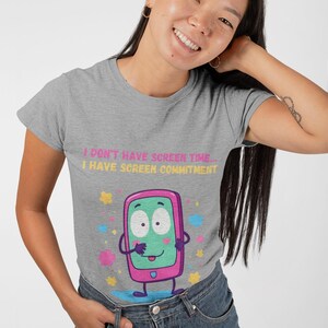 Może przedstawiać: Szara koszulka z napisem "I DON'T HAVE SCREEN TIME... I HAVE SCREEN COMMITMENT". Koszulka przedstawia kreskówkowy smartfon z zielonym ekranem i różową ramką. Kolorowe gwiazdki i kształty otaczają telefon.