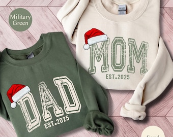 May include: Two crewneck sweatshirts, one military green and one sand-colored. The green sweatshirt says "DAD EST.2025" in white, with a Santa hat graphic. The sand sweatshirt says "MOM EST.2025" in green, also with a Santa hat. Both have a distressed, vintage look.