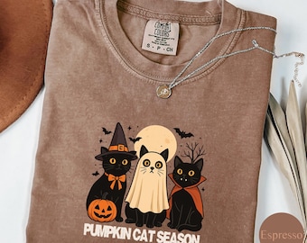 May include: Espresso-colored t-shirt featuring a Halloween-themed graphic with three black cats dressed as a witch, ghost, and vampire. The shirt includes the text "PUMPKIN CAT SEASON" and a Comfort Colors label.