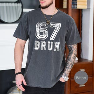 May include: Dark grey t-shirt with the number "67" and the word "BRUH" in a distressed white font. The shirt has a vintage, worn look. The shirt is a casual, everyday wear item.