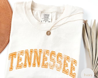 May include: Ivory-colored Comfort Colors t-shirt with the word "TENNESSEE" printed in orange block letters. A gold necklace with a pendant rests on the shirt. The shirt is folded on a white surface.
