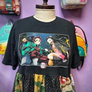 May include: Black t-shirt with a colorful graphic from "Demon Slayer" featuring two characters in action poses. The shirt has a layered design with a patterned skirt-like bottom. The text "Demon Slayer" is visible.