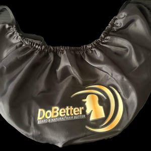 Puede incluir: Una gorra de barba negra con un logotipo dorado que dice "DoBetter BEARD & NATURAL HAIR BUTTER". La gorra tiene una banda elástica y está hecha de un material brillante y sedoso. El logotipo presenta una silueta de una persona con barba.