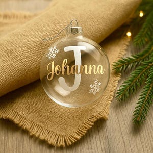 Könnte beinhalten: Transparente Glaskugel mit dem Namen "Johanna" in goldener Schrift und einem großen weißen "J". Zwei weiße Schneeflocken sind ebenfalls auf dem Ornament. Die Kugel hängt an einem silbernen Draht und liegt auf einem beigen Jutegewebe.