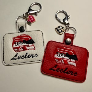 Könnte beinhalten: Zwei Schlüsselanhänger, einer weiß und einer rot, jeweils mit einem Rennhelm-Design mit der Nummer 16 und dem Namen "Leclerc". Die Schlüsselanhänger haben silberne Verschlüsse und herzförmige Anhänger.