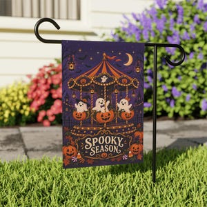 May include: A rectangular garden flag hanging on a black metal stand, featuring a dark purple illustration of a carousel with ghosts riding in pumpkin seats. The text "SPOOKY SEASON" is printed in a decorative font at the bottom with pumpkins.