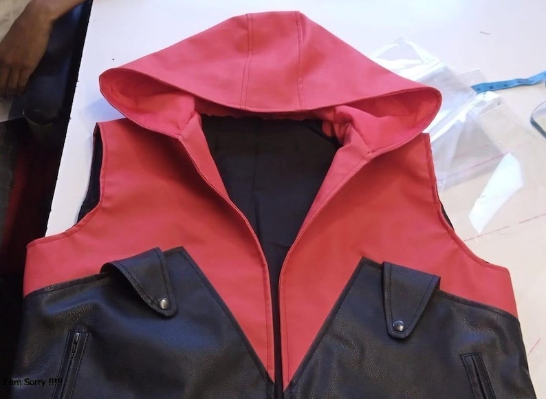 May include: A red and black hooded vest with a black interior. The vest features a red hood and upper section, with black panels and pockets. The pockets have a flap and a silver button. The vest is likely made of a leather-like material.