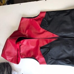 May include: A two-tone vest in red and black. The vest features a red front and hood, with black panels on the sides and back. It has a zipper closure and small pockets. The garment is made of a smooth, possibly leather-like material.