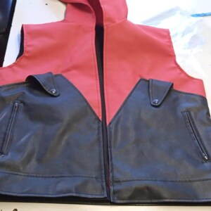 May include: A red and black faux leather vest with a hood. The vest has a black front with two pockets and a zipper closure. The top of the vest and hood are red. The vest is laying flat on a white surface.