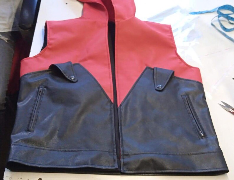 May include: A red and black faux leather vest with a hood. The vest has a black front with two pockets and a zipper closure. The top of the vest and hood are red. The vest is laying flat on a white surface.