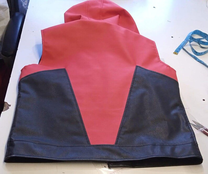 May include: A red and black vest with a hood. The vest features a red body with black panels on the sides and bottom. The hood is also red. The vest is made of a smooth material and appears to be new.