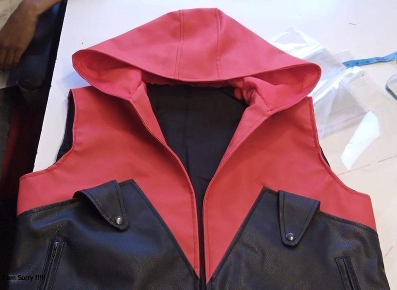May include: A red and black hooded vest with two front pockets. The vest features a red hood and upper section, with a black lower section and pockets. The vest is made of a smooth material and has a zipper.