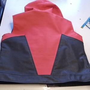 May include: A red and black vest with a hood. The back of the vest is red with black panels on the lower portion. The vest is made of a leather-like material. A blue measuring tape and scissors are on the table.