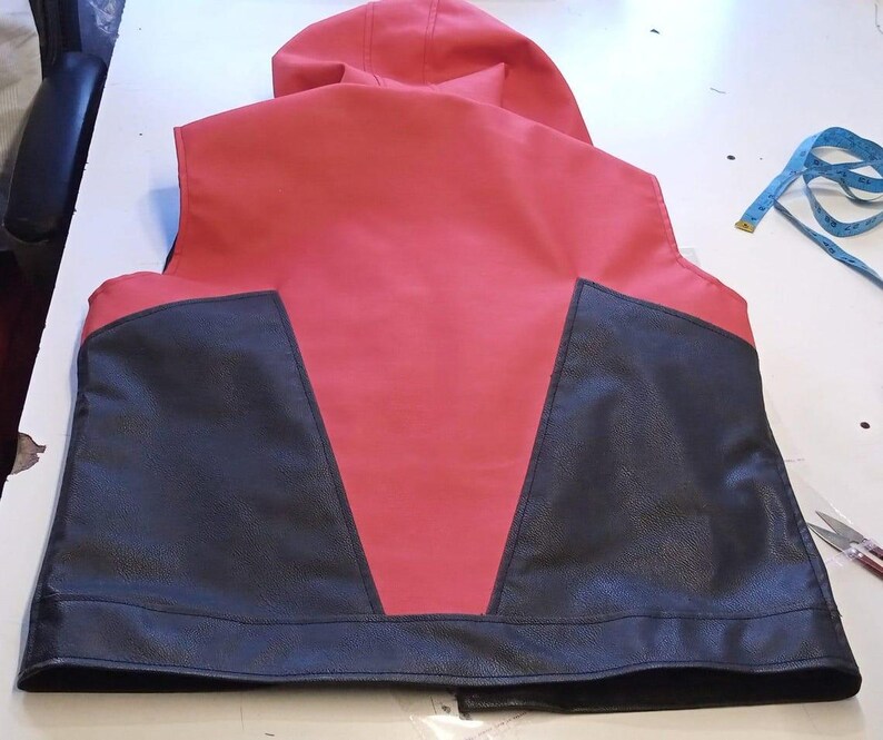 May include: A red and black vest with a hood. The back of the vest is red with black panels on the lower portion. The vest is made of a leather-like material. A blue measuring tape and scissors are on the table.