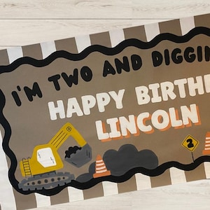 May include: A banner with the text "I'M TWO AND DIGGING IT! HAPPY BIRTHDAY LINCOLN" in white and orange lettering. The banner features illustrations of construction vehicles, including a yellow excavator and a dump truck, with orange traffic cones.