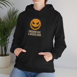 May include: Black hooded sweatshirt with a front pocket. The sweatshirt features a graphic of a yellow, evil-looking smiley face with the text "FREEDOM HAS A WICKED GRIN" in white. The hoodie is made of a soft material.