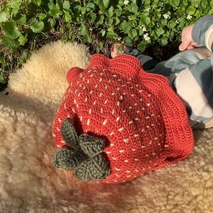 Könnte beinhalten: Eine handgefertigte, rote gehäkelte Erdbeere mit weißen Samendetails und grünen Blättern. Die Erdbeere liegt auf einer cremefarbenen, strukturierten Oberfläche, mit grünem Laub im Hintergrund.