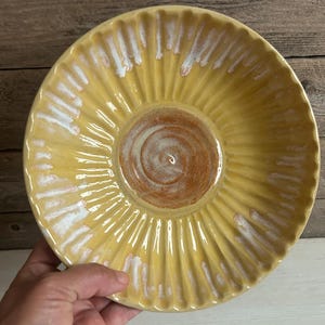 May include: A round ceramic bowl with a fluted, scalloped edge. The interior features a bright yellow glaze with white drip accents along the ridges, while the center displays a circular, unglazed terracotta-colored spiral pattern.